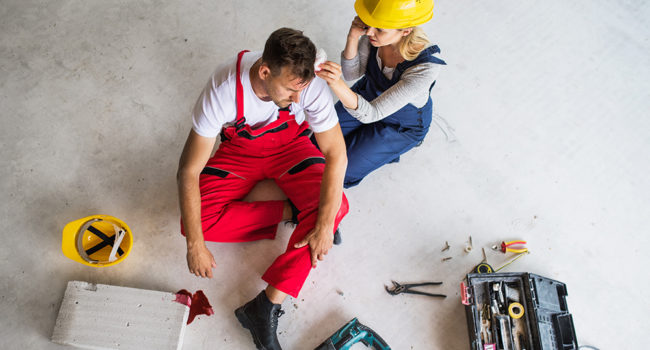 Accident of a male worker at the construction site. A woman with smartphone helping her injured colleague, wiping his forehead. Top view.